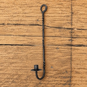 Pode incluir: Um castiçal de parede em metal preto com uma haste vertical longa e fina de textura torcida e um anel circular para pendurar no topo. A base consiste num pequeno prato circular plano com um copo cilíndrico central.