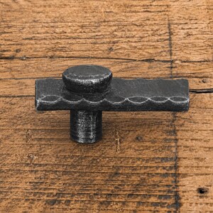 Könnte beinhalten: Ein dunkelgrauer Möbelknopf aus Metall mit einer horizontalen, rechteckigen Stange mit gewellter Kante und einem kreisförmigen Detail oben. Der Beschlag hat eine texturierte, handgeschmiedete Optik mit einem zylindrischen Sockel.