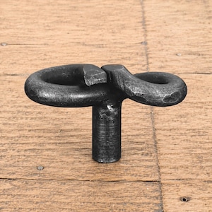 May include: A dark grey metal cabinet knob featuring a T-shaped base topped with two interconnected, rounded loops that form a horizontal figure-eight shape. The surface of the metal has a textured, matte finish with visible hammer marks.