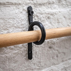 May include: A black metal bracket with a circular ring holds a wooden dowel against a white brick wall. The bracket is secured with a single screw. The dowel is a light brown color.