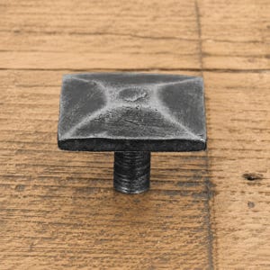 May include: A square cabinet knob with a dark grey, textured, metallic finish. The knob features a slightly raised, pyramid-like centre and a cylindrical base with a threaded post, showcasing a hammered, industrial appearance with visible surface details.