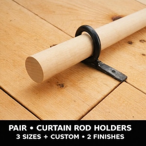 Puede incluir: Una barra de cortina de madera de color marrón claro sujeta por un soporte de metal negro. La barra es cilíndrica. El soporte es de color negro mate oscuro. La imagen está sobre una superficie de madera marrón claro. Texto en la parte inferior: "PAIR • CURTAIN ROD HOLDERS".