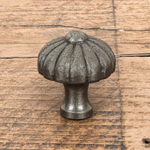 Peut inclure: Un bouton de meuble en métal gris foncé avec une tête en forme de champignon présentant des cannelures verticales et une finition texturée et patinée. Le bouton possède une base cylindrique lisse qui s'affine légèrement vers le centre.