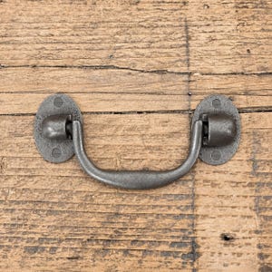 Peut inclure: Une poignée tombante en métal gris foncé composée d'une prise incurvée fixée à deux plaques de montage ovales. Chaque plaque présente deux trous de vis visibles, et la quincaillerie affiche une finition mate texturée au design simple.