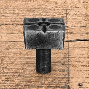 May include: A dark grey, square-shaped metal knob with a textured, weathered finish. The top surface features a cross-shaped indentation with four circular depressions in each corner. A cylindrical base extends downwards from the centre of the square top.
