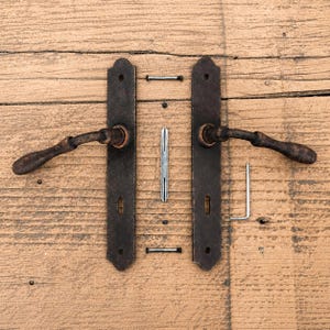 May include: A set of two dark brown, textured metal door handles with rectangular backplates, each featuring a curved lever. The set includes a central square spindle, two small mounting screws, and an L-shaped metal hex key tool for assembly.