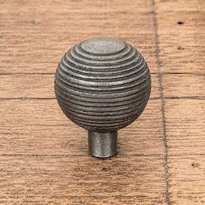 Puede incluir: Un pomo esférico para muebles con un acabado metálico gris oscuro y una serie de estrías horizontales y concéntricas que rodean toda la superficie. El pomo se apoya sobre una base cilíndrica corta que sale del centro inferior.