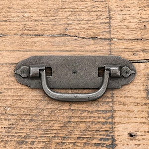May include: A dark grey metal drawer pull with a rectangular backplate featuring rounded ends and decorative pointed edges. The handle is a U-shaped drop pull attached to the plate with two hinges. Three visible Phillips head screws secure the hardware.