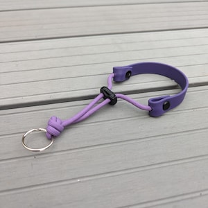 May include: A purple key fob with a matching cord and a silver key ring. The fob is made of a flexible material and has a black plastic adjuster. The cord is knotted to secure the key ring.