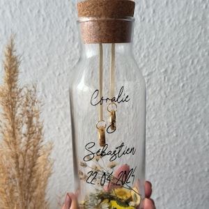 Peut inclure: Une bouteille en verre transparente avec un bouchon en liège remplie de fleurs séchées et deux anneaux dorés suspendus à une ficelle. La bouteille est étiquetée avec les noms "Coralie" et "Sébastien" et la date "22.04.2023".