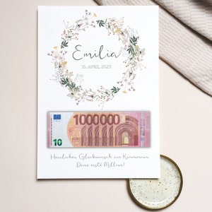 May include: A white card with a floral wreath and the text "Emilia 16. APRIL 2023". A fake 10 euro bill with the number 1,000,000 printed on it is attached to the card. The text "Herzlichen Gl&uuml;ckwunsch zur Kommunion Deine erste Million!" is printed below the bill.
