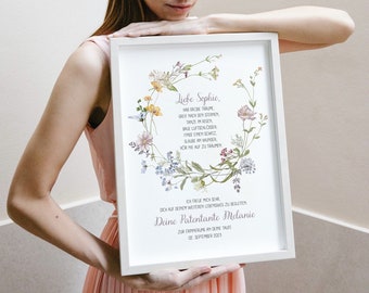 May include: A framed print with a white border, featuring a floral wreath design and German text. The text includes the words "Liebe Sophie" and "Deine Patentante Melanie". The frame is held by a person.