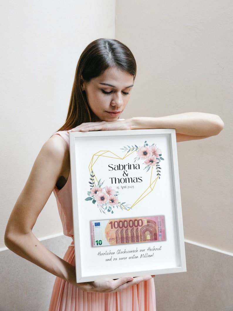 May include: A white frame with a gold heart design and floral accents. The text "Sabrina & Thomas 15. April 2023" is printed on the frame. A 1,000,000 euro bill is displayed inside the frame with the text "Herzlichen Gl&uuml;ckwunsch zur Hochzeit und zu eurer ersten Million!"