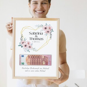 May include: A framed wedding gift card with a floral design and a gold geometric heart. The card includes the names Sabrina & Thomas, the date 15. April 2023, and a fake 1 million euro bill. The text reads "Herzlichen Gl&uuml;ckwunsch zur Hochzeit und zu eurer ersten Million!"
