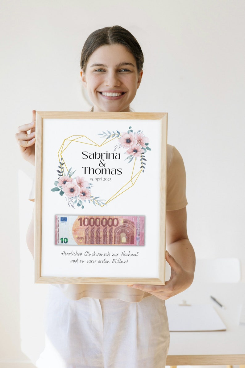 May include: A framed wedding gift card with a floral design and a gold geometric heart. The card includes the names Sabrina & Thomas, the date 15. April 2023, and a fake 1 million euro bill. The text reads "Herzlichen Gl&uuml;ckwunsch zur Hochzeit und zu eurer ersten Million!"