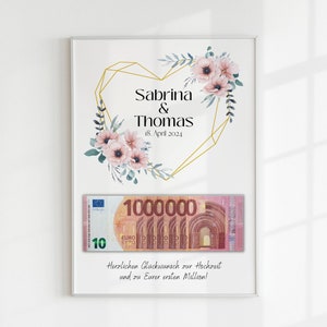 May include: A white framed print with a gold geometric heart design and pink flowers. The text "Sabrina & Thomas 18. April 2024" is inside the heart. A 1,000,000 euro bill is displayed below the heart with the text "Herzlichen Gl&uuml;ckwunsch zur Hochzeit und zu Eurer ersten Million!"