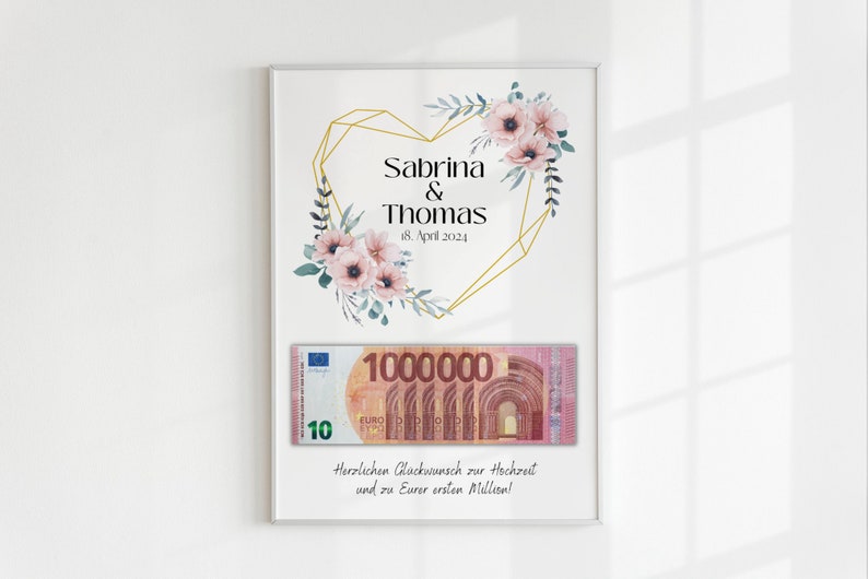 May include: A white framed print with a gold geometric heart design and pink flowers. The text "Sabrina & Thomas 18. April 2024" is inside the heart. A 1,000,000 euro bill is displayed below the heart with the text "Herzlichen Gl&uuml;ckwunsch zur Hochzeit und zu Eurer ersten Million!"