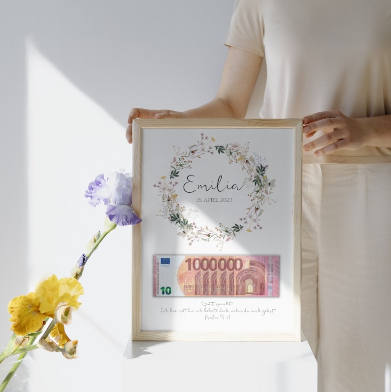 May include: A framed print with a floral wreath design and the name "Emilia" and the date "16. APRIL 2023". A 100,000 Euro bill is placed inside the frame. The text "Gott segnet dich" and "Ich bin mit dir und beh&uuml;te dich, wohin du auch gehst" is printed below the bill.