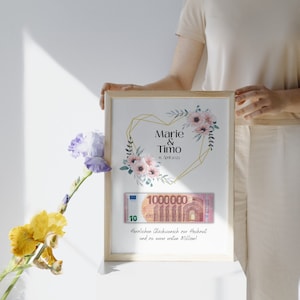 May include: A framed print with a gold geometric heart design and pink flowers. The print says "Marie & Timo" and "15. April 2023". A fake 10 euro bill with the number 1000000 is inside the frame. The text below the heart says "Herzlichen Gl&uuml;ckwunsch zur Hochzeit und zu eurer ersten Million!"