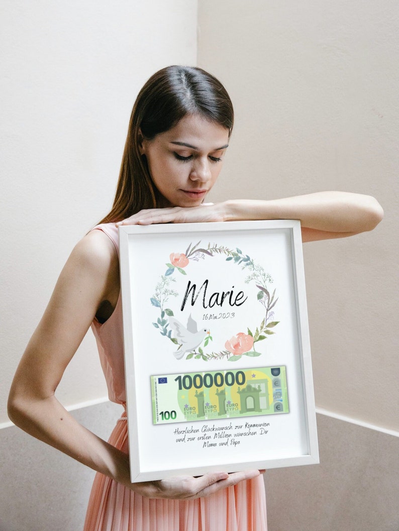 May include: A white framed print with a floral wreath and a dove. The print has a fake 100 euro bill with the number 1,000,000 printed on it. The text on the print reads "Marie 16. Mai 2023 Herzlichen Gl&uuml;ckwunsch zur Kommunion und zur ersten Million w&uuml;nschen Dir Mama und Papa."