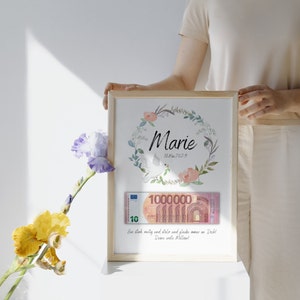 May include: A framed print with a floral wreath and the name "Marie" in cursive. The date "16.Mai.2023" is written below the name. A 10 euro bill is visible in the frame, with the text "See stark, neitig und stelo und gloube wour on Dich! Deme erste Mellow!" below the bill.