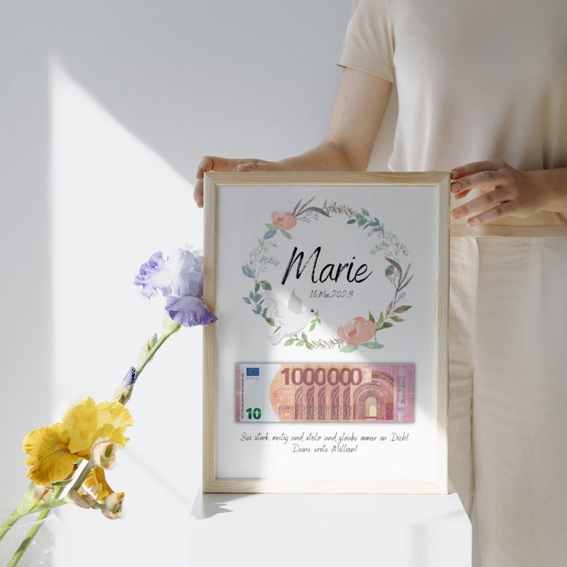 May include: A framed print with a floral wreath and the name "Marie" in cursive. The date "16.Mai.2023" is written below the name. A 10 euro bill is visible in the frame, with the text "See stark, neitig und stelo und gloube wour on Dich! Deme erste Mellow!" below the bill.