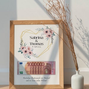 May include: A framed print with a gold geometric heart design and floral accents. The text reads "Sabrina & Thomas 18. April 2024". A 10 Euro bill with the text "1000000" is visible below the heart. The text below the bill reads "Herzlichen Gl&uuml;ckwunsch zur Hochzeit und zu Eurer ersten Million!"