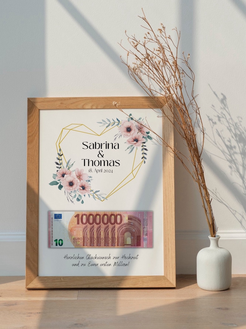 May include: A framed print with a gold geometric heart design and floral accents. The text reads "Sabrina & Thomas 18. April 2024". A 10 Euro bill with the text "1000000" is visible below the heart. The text below the bill reads "Herzlichen Gl&uuml;ckwunsch zur Hochzeit und zu Eurer ersten Million!"
