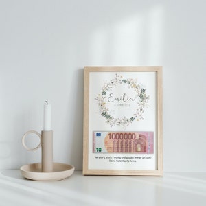 May include: A framed print with a floral wreath design and the name "Emilia" and the date "10 APRIL 2023". The print also includes a 10 Euro bill with the text "Sei stark, stolz & mutig und glaube immer an Dich! Deine Patentante Anna."