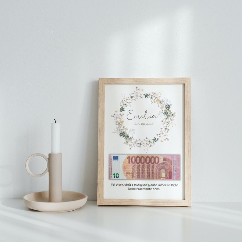 May include: A framed print with a floral wreath design and the name "Emilia" and the date "10 APRIL 2023". The print also includes a 10 Euro bill with the text "Sei stark, stolz & mutig und glaube immer an Dich! Deine Patentante Anna."