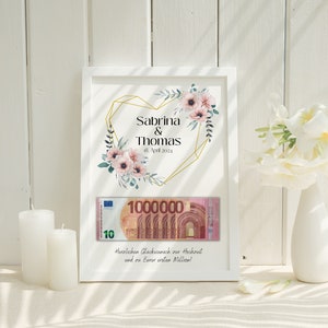 May include: A white frame with a gold heart design and floral accents. The text "Sabrina & Thomas 18. April 2024" is printed on the frame. A 10 Euro bill is visible inside the frame. The text "Herzlichen Gl&uuml;ckwunsch zur Hochzeit und zu Eurer ersten Million!" is printed below the bill.
