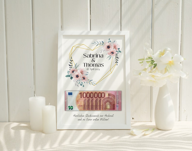 May include: A white frame with a gold heart design and floral accents. The text "Sabrina & Thomas 18. April 2024" is printed on the frame. A 10 Euro bill is visible inside the frame. The text "Herzlichen Gl&uuml;ckwunsch zur Hochzeit und zu Eurer ersten Million!" is printed below the bill.