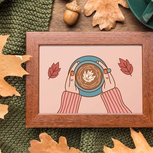 May include: A framed illustration of hands holding a blue mug with a leaf design on a pink background. The frame is brown, and the image is surrounded by fall leaves and a green knitted blanket. A cup of coffee is visible.