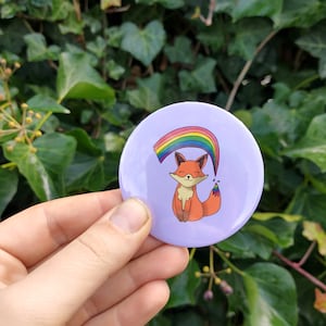 May include: A purple button with a rainbow and a cartoon fox. The fox is orange and has a white belly. The rainbow is above the fox and has red, orange, yellow, green, blue, indigo, and violet stripes.