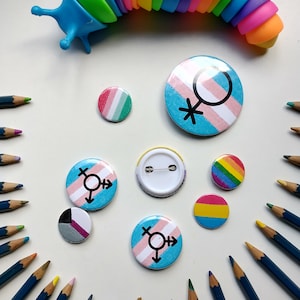 Peut inclure: Une collection d'épingles colorées avec différents drapeaux et symboles de la fierté LGBTQ+. Les épingles sont disposées sur une surface blanche avec un jouet arc-en-ciel en arrière-plan.
