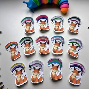 Op de afbeelding: Twaalf vossen stickers met verschillende pride-vlaggen. Elke sticker toont een cartoonvos met een regenboog-, bi-, transgender-, non-binair-, panseksueel-, lesbisch-, genderfluid-, aseksueel-, intersekse- en polyamoreuze pride-vlag.