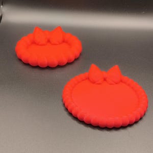 May include: Two bright red circular trays with a raised, scalloped edge and a decorative bow detail at the top. The trays have a smooth, flat interior surface and a consistent matte finish, designed as small containers for holding various small items.