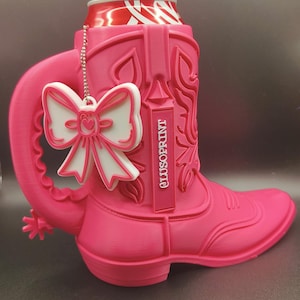 Könnte beinhalten: Ein leuchtend pinker Dosenhalter in Form eines Cowboystiefels mit Griff und Sporen. Der Stiefel zeigt geprägte Western-Muster und den Schriftzug CLUSOPRINT. Ein weiß-rosa Schleifenanhänger hängt an einer silbernen Kette am Griff.
