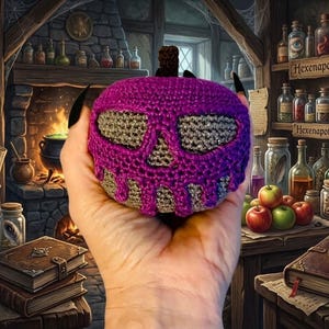 Peut inclure: Objet en forme de crâne crocheté violet et gris. Le motif de crâne est visible sur le devant, avec les yeux et le nez en gris. L'objet est tenu dans une main, avec un arrière-plan d'apothicairerie de sorcière.