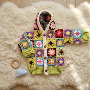Puede incluir: Un cárdigan de bebé de ganchillo hecho a mano y colorido con capucha. El suéter presenta un diseño de patchwork con cuadrados en varios colores, incluyendo amarillo, morado, rosa y verde. Tiene una parte delantera abotonada y está sobre una superficie blanca y esponjosa.
