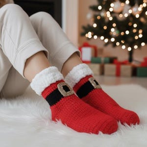 May include: Red crochet socks with a Santa Claus design. The socks feature a black band with a tan buckle and a white cuff. The socks are on a white fluffy surface, with a Christmas tree and gifts in the background.