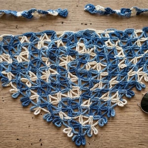 May include: A handmade, blue and white crochet shawl with a floral pattern. The shawl is triangular and has a scalloped edge. Sunglasses and a brown case are next to the shawl on a wooden surface.