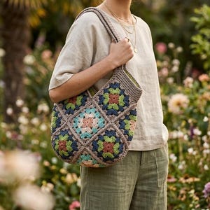May include: A crocheted tote bag with a floral pattern in shades of blue, green, orange, and pink. The bag is held by a person wearing a beige top and olive green pants. The background is a garden.