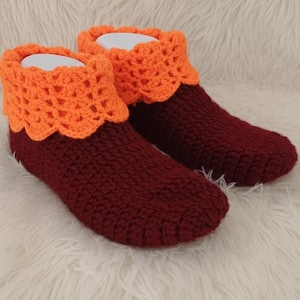 May include: A pair of handmade crochet slippers. The slippers are primarily burgundy with orange cuffs. The cuffs have a scalloped edge. The slippers are displayed on a white, fluffy surface.