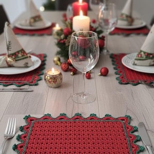 May include: A Christmas table setting with red crocheted placemats, green scalloped edges, white plates, napkins, candles, and wine glasses. The scene evokes a cosy festive atmosphere, perfect for a holiday meal.