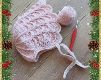 May include: A handmade, light pink crochet baby bonnet with a scalloped edge and tie strings. A ball of matching yarn and a red crochet hook are next to the bonnet. The bonnet is on a wooden surface with a Christmas-themed border.