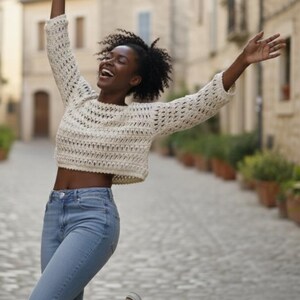 May include: A cream-colored, open-weave crochet sweater with long sleeves. The sweater is paired with light blue denim jeans. The person is smiling and has their arms raised in the air.