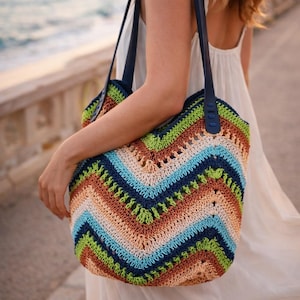 May include: A colorful crochet tote bag with a chevron pattern in shades of blue, green, orange, and cream. The bag has dark blue straps and is held by a person wearing a white dress. The bag is a handmade item.