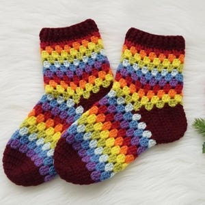 Puede incluir: Un par de calcetines de ganchillo hechos a mano. Los calcetines presentan un patrón de arcoíris con cuadrados rojos, naranjas, amarillos, verdes, azules y morados. El talón y el puño son de color burdeos oscuro. Los calcetines se muestran sobre una superficie blanca y esponjosa.