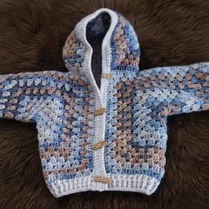 May include: A hooded cardigan featuring a granny square crochet pattern in shades of blue, tan, and white. The garment includes long sleeves, a white crocheted border along the front opening and hood, and three light-colored wooden toggle buttons.